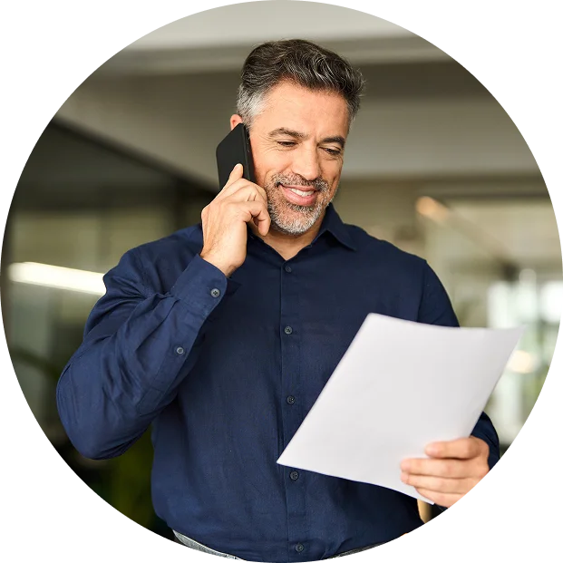 A man with gray hair and a beard, wearing a navy blue shirt, is talking on the phone while reading a document.