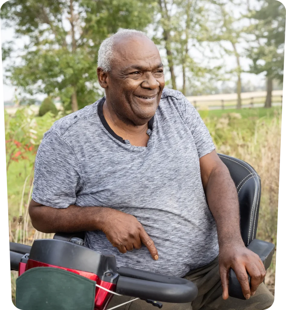 An older man with gray hair sits on a mobility scooter outdoors, smiling