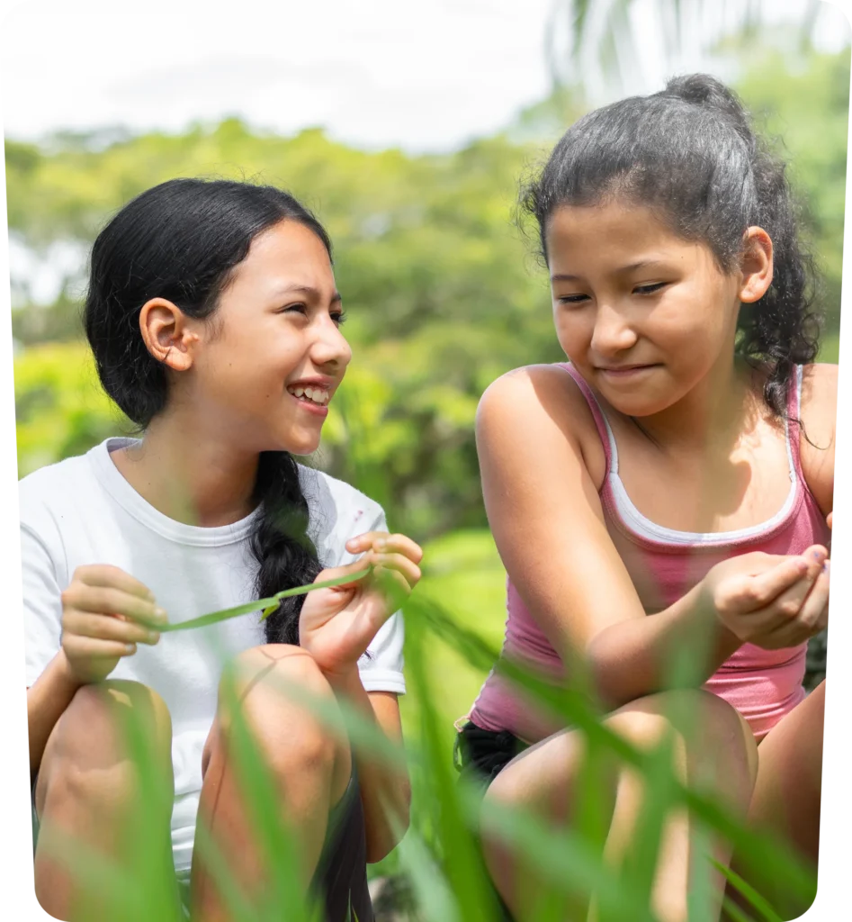 Two girls sit outdoors in a grassy area, smiling and holding blades of grass, with trees blurred in the background.
