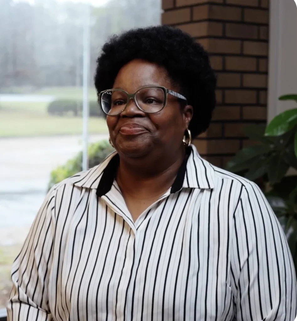 A woman with short curly hair, glasses, and striped shirt stands indoors, smiling